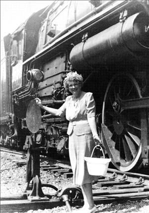 A woman stands next to a steam locomotive holding a purse and wearing a skirt suit and pumps. 